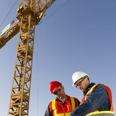 Two construction workers in hard hats and safety vests discuss plans under a crane.
